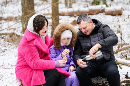 Family with child sits on fallen tree during rest stop, drinking hot tea from thermos with cookies....