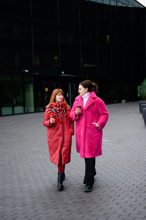 Two women are drinking coffee while walking down city street, chatting animatedly. Girlfriends are talking and laughing in winter urban setting, looking at each other. Friendship and communication.の写真素材