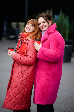 Portrait of girlfriends enjoying winter coffee, leaning on each other and smiling happily. Women laugh, share joyful moments over cup of coffee on cold winter days. Concept of friendship, coffee breakの写真素材