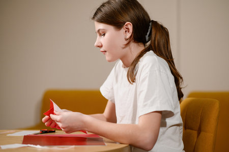 Girl in white shirt carefully folds origami while sitting at table in living room. Student with hair pulled back in ponytail intently folds paper into shape of bird. Childrens art concept.の写真素材