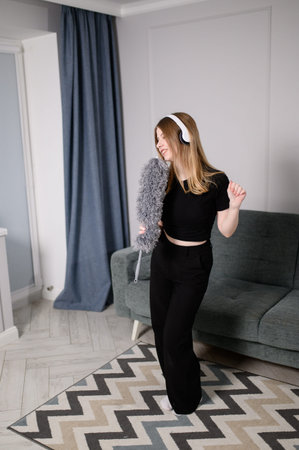 Caucasian woman dances in living room with headphones, listening to music while cleaning and...