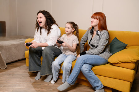 Happy family spends free time playing video games. Mom and daughter play with video game controller while sitting on living room sofa, enjoying eSports, competing, having fun while playing video gamesの写真素材