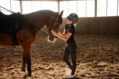 Woman wearing riding harness, adjusts horses bridle in indoor arena, caring for beloved pet. Rider strokes horse with hand, owner taking care of animal. Concept: equestrian sport, riding school.の写真素材
