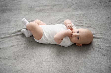 Cute white baby lies on its back, sucking its fingers, on soft gray blanket. Infant self-discovery....