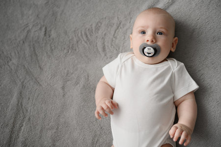 Calm baby looks up, newborn lies on bed in nursery with pacifier in his mouth. Baby concept, baby...