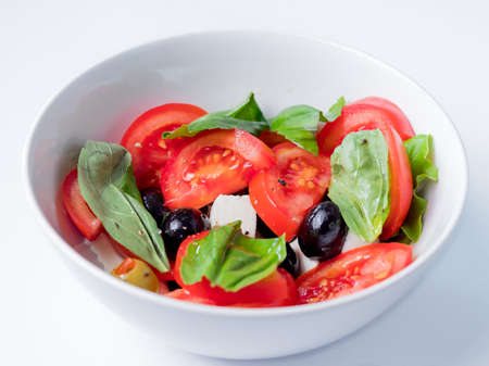 Close up photo of Italian mediterranean salad with tomatoes, olives, mozzarella cheese, basil and olive oil dressing in a white bowlの写真素材