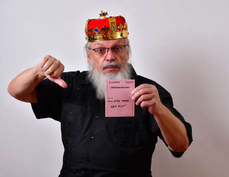 Tyrannical boss humiliates an employee with a nasty memo firing him or her.  Mature jerk with a golden crown and black guayabera shirt and long white beard.の写真素材