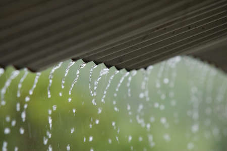 Streams of rain water pour off a corrigated roof, grey roof, green background selective focusの写真素材