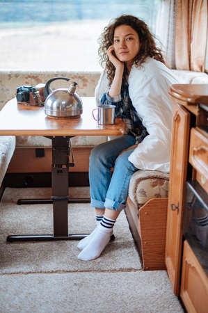 Girl sitting at a table in a motorhome in a white blanket and holding a mugの写真素材