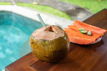 Open coconut with glass straw and sliced papaya near swimming poolの写真素材