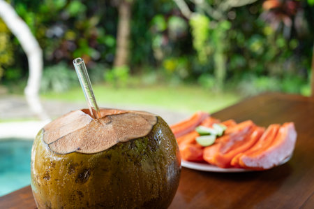 Open coconut with glass straw and sliced papaya near swimming poolの写真素材