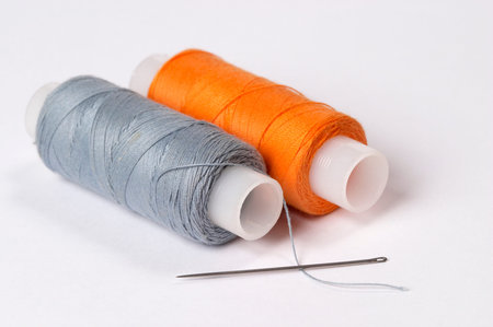 Two coils of thread grey and orange with needle over white background