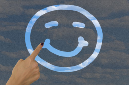 hand drawing a smiley face on a dirty window with a finger revealing clear blue sky on the other...