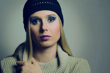  fashion portrait of a blonde woman in studio wearing a heavy make up and a turtleneck sweater with a black knitted hatの写真素材