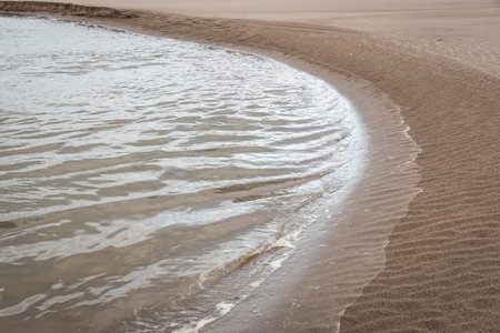 Small, brightly shimmering waves lapping against the curving sweep of a sandy beach shoreline, St...
