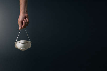 front view of single male hand carrying corona virus protection mask by the elastic, set against dramatic dark background and lightingの写真素材