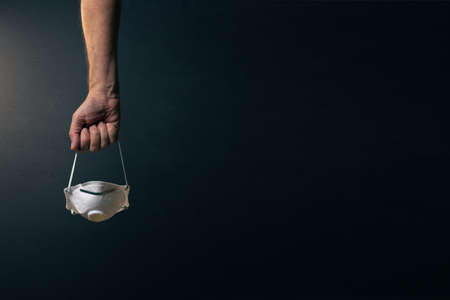 front view of single male hand clenched carrying corona virus protection mask by the elastic, set against dramatic dark background and lightingの写真素材