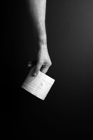 black & white image of single male hand carrying toilet roll by finger tips, set against dramatic...