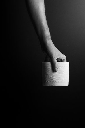 black & white image of single male hand carrying toilet roll by finger tips, set against dramatic dark background and lightingの写真素材