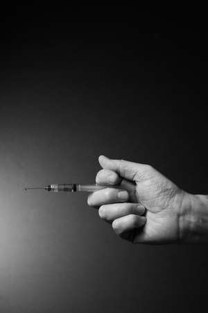 Black and white image of man's clenched hand holding syringe horizontally with liquid droplet on needle point, with dramatic lightingの写真素材