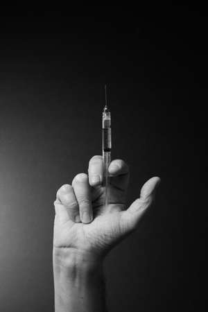 Black and white image of man's open hand holding syringe vertically upwards with liquid droplet on needle point, with dramatic lightingの写真素材