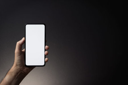 Colour image of man's hand holding black smartphone vertically with blank white screen isolated on...
