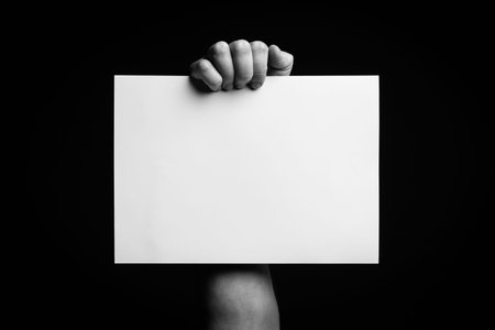 B+W image of male hand holding blank sheet of paper against black background with space for copy.