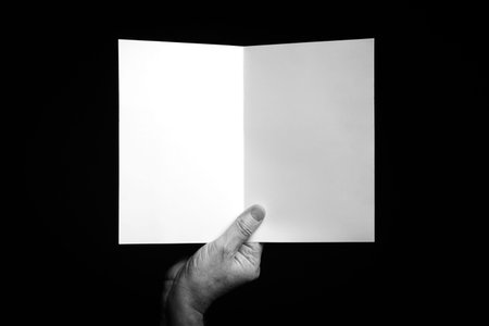 B+W image of male hand holding folded blank sheet of paper or card against black background with space for copy.の写真素材