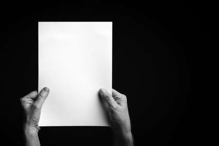 B+W image of male hands holding blank sheet of paper against black background with space for copy.の写真素材