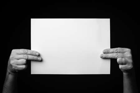 B+W image of male hands holding blank sheet of paper against black background with space for copy.の写真素材