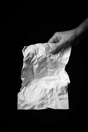B+W image of male hand holding crumpled sheet of paper against black background with dramatic lighting.の写真素材