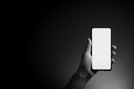 Black and white image of man's hand holding black smartphone vertically with blank white screen...