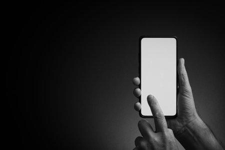 Black and white image of man's hand holding black smartphone and using blank white screen with...