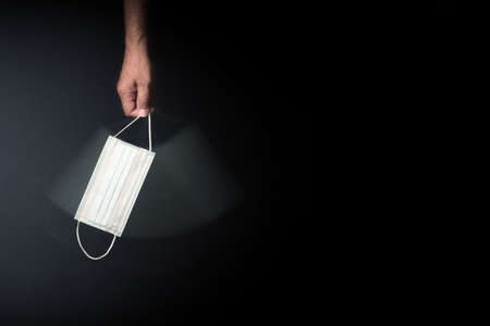 male hand holding & swinging single medical mask against dark background with copy spaceの写真素材