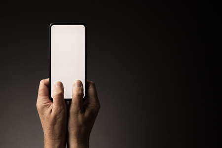 Man's hands holding smart phone and texting with blank white screen isolated on dark background with copy spaceの写真素材