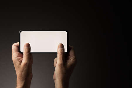Man's hands holding smart phone horizontally texting or gaming with blank white screen isolated on dark background with copy spaceの写真素材