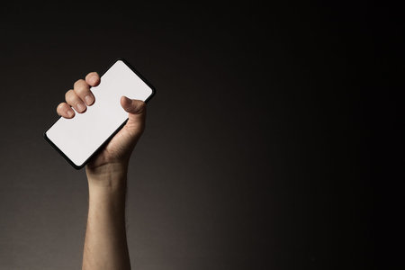 Man's hand holding smart phone with blank white screen isolated on dark background with copy space