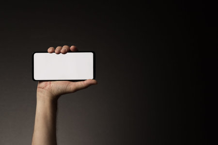 Man's hand holding smart phone horizontally with blank white screen isolated on dark background with copy spaceの写真素材
