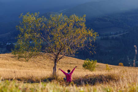 Young girl rises up her hands while sitting under tree an mountain backgroundの写真素材