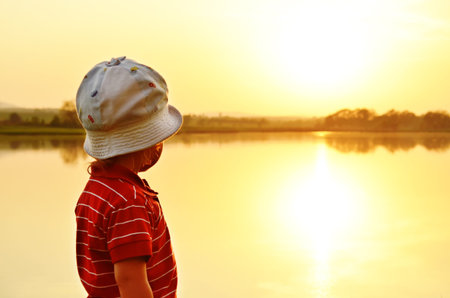 A little boy stands on the shore of the lake and watching the sunset  Orange background の写真素材
