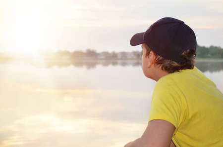 Boy in a baseball cap sits on the shore of the lake and looking at the sunsetの写真素材