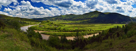 Panorama with highest point - green mountains and river, shined with the sun. Fluffy clouds in the blue sky.の写真素材