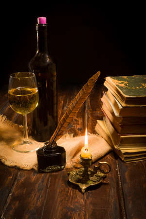 Concept of education and writing, an inkwell with a pen and a lit candle on the background of a book scroll and a bottle of wine with a glassの写真素材