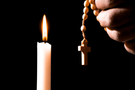 Lit white candle and a hand holding a rosary, isolated on a black background