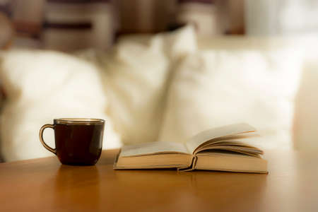 A glass of coffee and a book on the table, on a background of pillows on a light bedの写真素材
