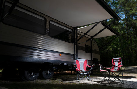 Red camping chairs under the awning of a travel trailer in a forested campsiteの写真素材