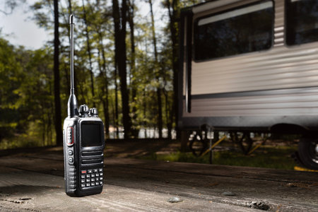 Small walkie talkie ideal for the outdoors on a picnic table with a large travel trailer parked in a forest backlit by the sun while camping near a lake that can be seen through the trees glistening below.の写真素材