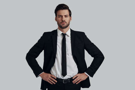 Portrait of confidence. Handsome young man in full suit looking at camera and keeping hands on hips while standing against grey backgroundの写真素材