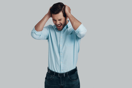 Failure. Frustrated young man keeping head in hands and making a face while standing against grey...