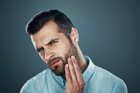 Need to shave. Handsome young man touching his beard and looking at camera while standing against grey backgroundの写真素材